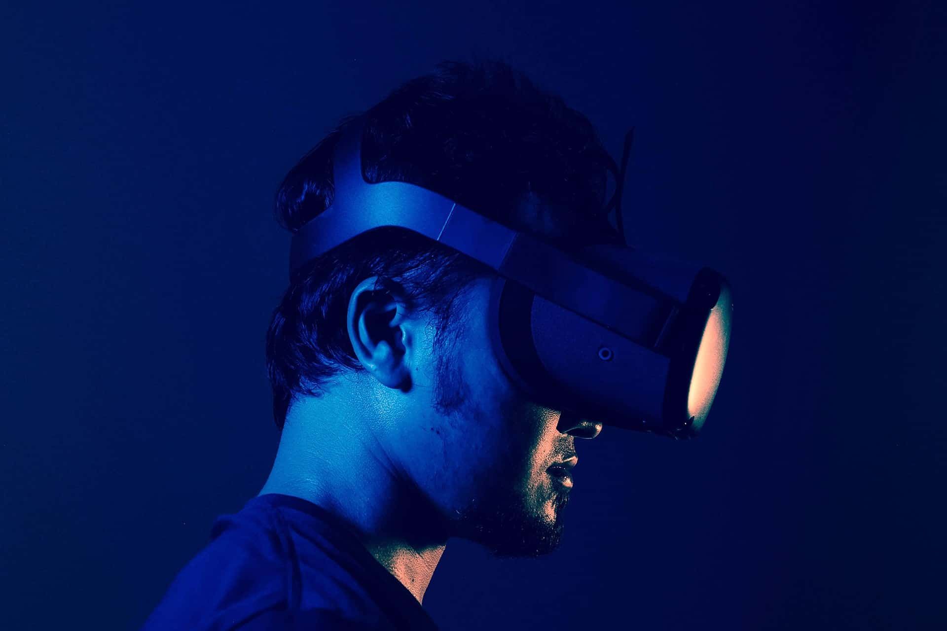 A person wears a virtual reality headset, illuminated by blue and orange light, against a dark background.