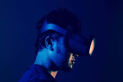 A person wears a virtual reality headset, illuminated by blue and orange light, against a dark background.